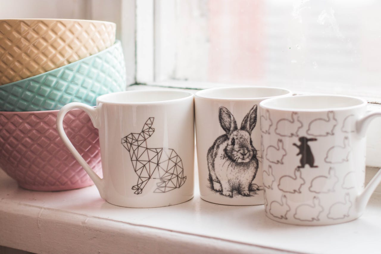 Elegant ceramic mugs with rabbit designs and colorful bowls on a windowsill, perfect for home decor inspiration.
