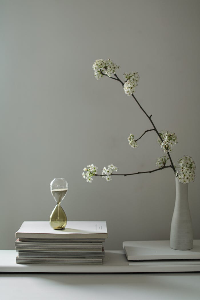 A minimalist still life featuring a floral branch, hourglass, and stacked books.